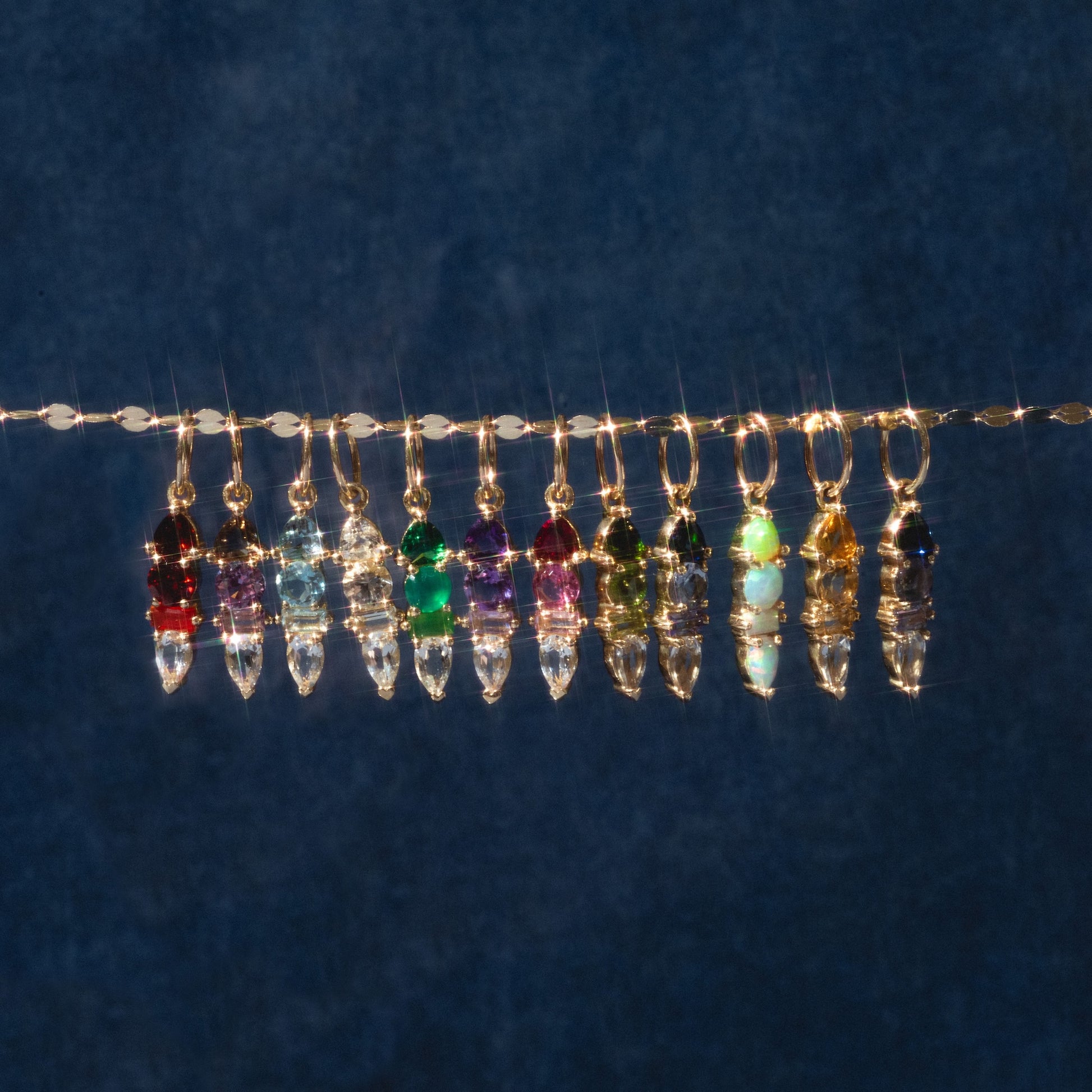 Twelve Solid Gold Modern Ombre Birthstone Charms with gold loops hang on a thin wire against a dark blue background, arranged in a sparkling, colorful spectrum.