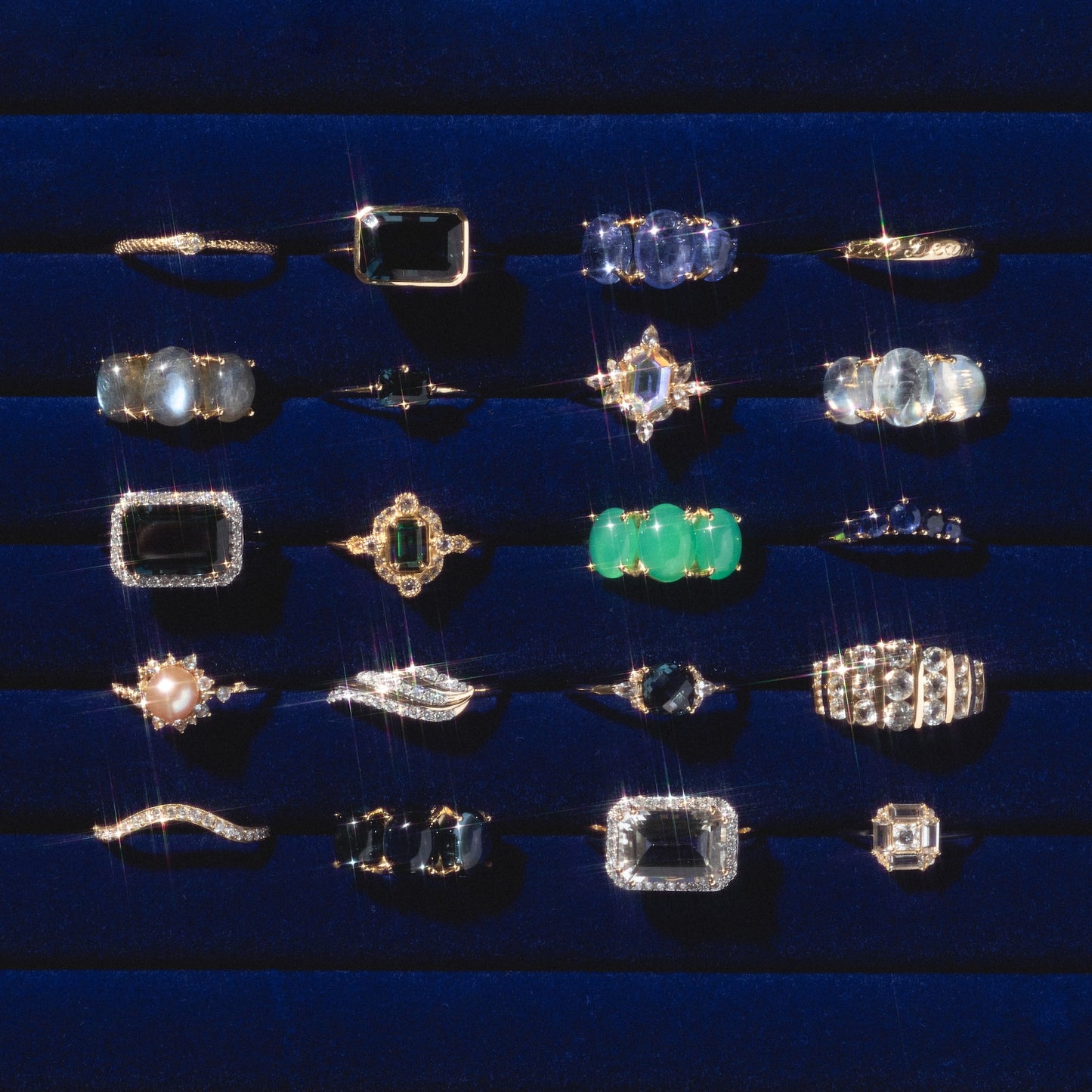 A selection of 20 gemstone rings, including the 14k Till Death Layering Ring and standout Charlie and Marcelle designs, displayed on a dark blue velvet tray in five neat rows with gems of various shapes, sizes, and colors.