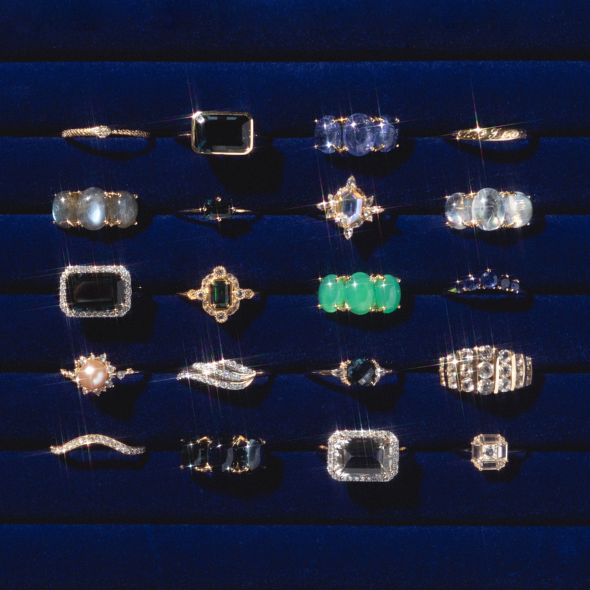 A selection of 20 gemstone rings, including the 14k Till Death Layering Ring and standout Charlie and Marcelle designs, displayed on a dark blue velvet tray in five neat rows with gems of various shapes, sizes, and colors.