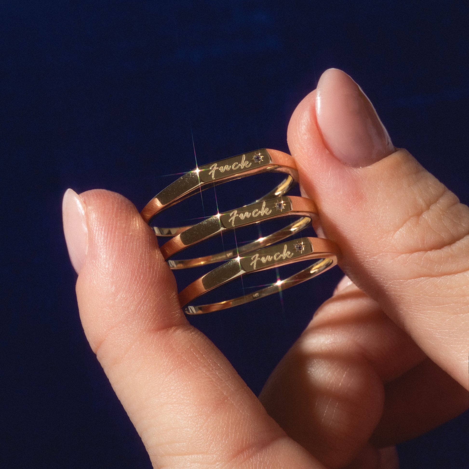 A manicured hand displays three Solid Gold Tiny Fuck Rings, each engraved with "Fuck" and a small star, set against a dark blue background.