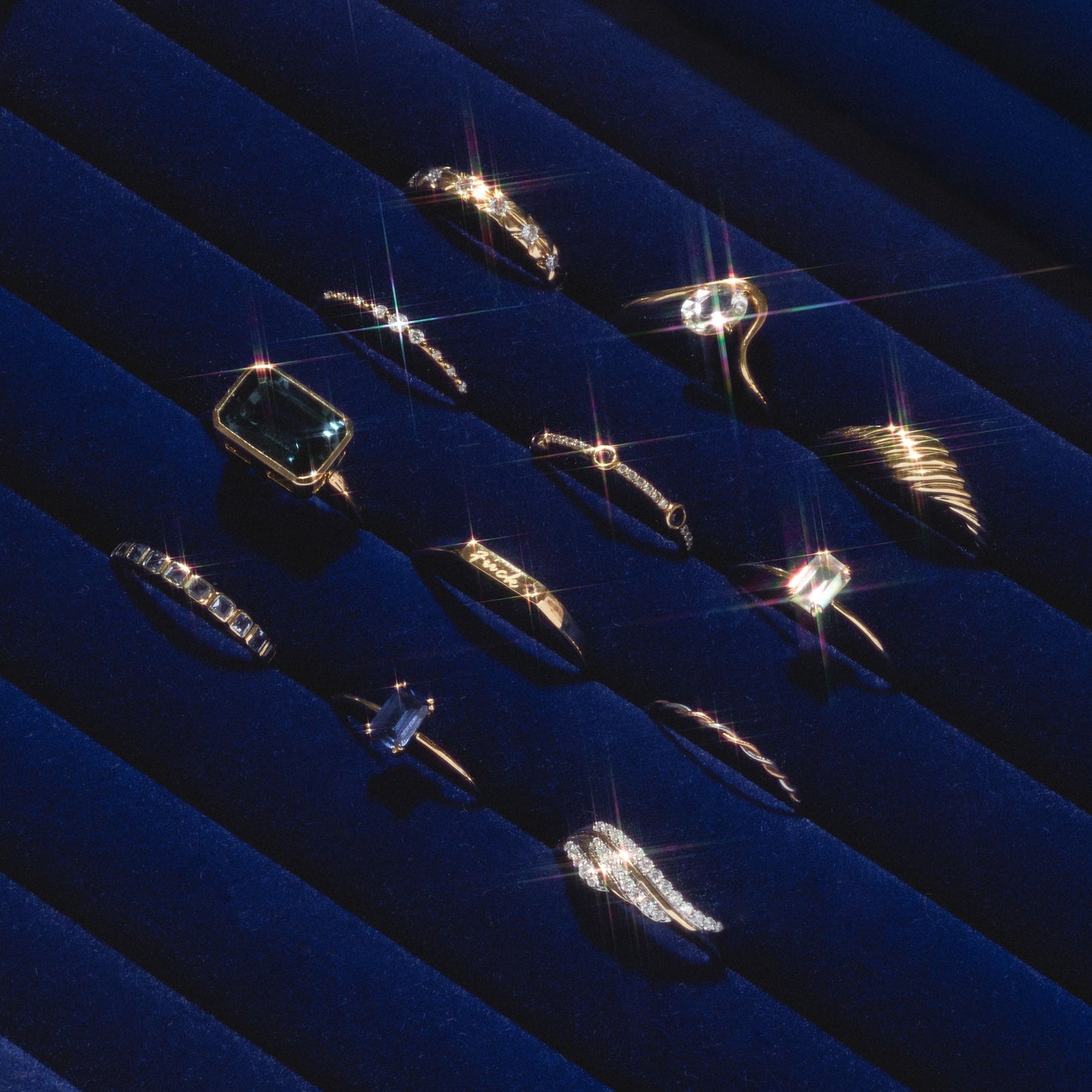 The 14k Solid Gold Josephine Diamond Crown Ring is displayed among gold and silver rings on dark blue velvet rolls, sparkling under the light and casting star-like reflections across their glossy surfaces.