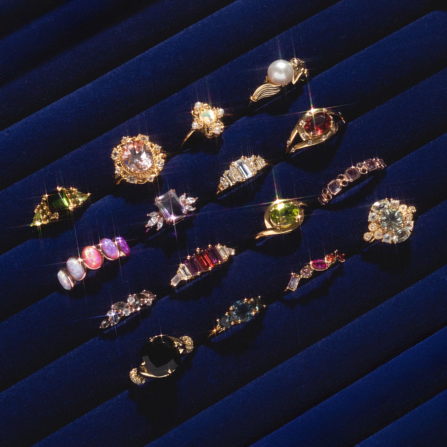 The Past Life Black Spinel Statement Ring shines among vintage rings with colorful gemstones and intricate gold settings, all displayed in neat rows on a dark blue velvet background.