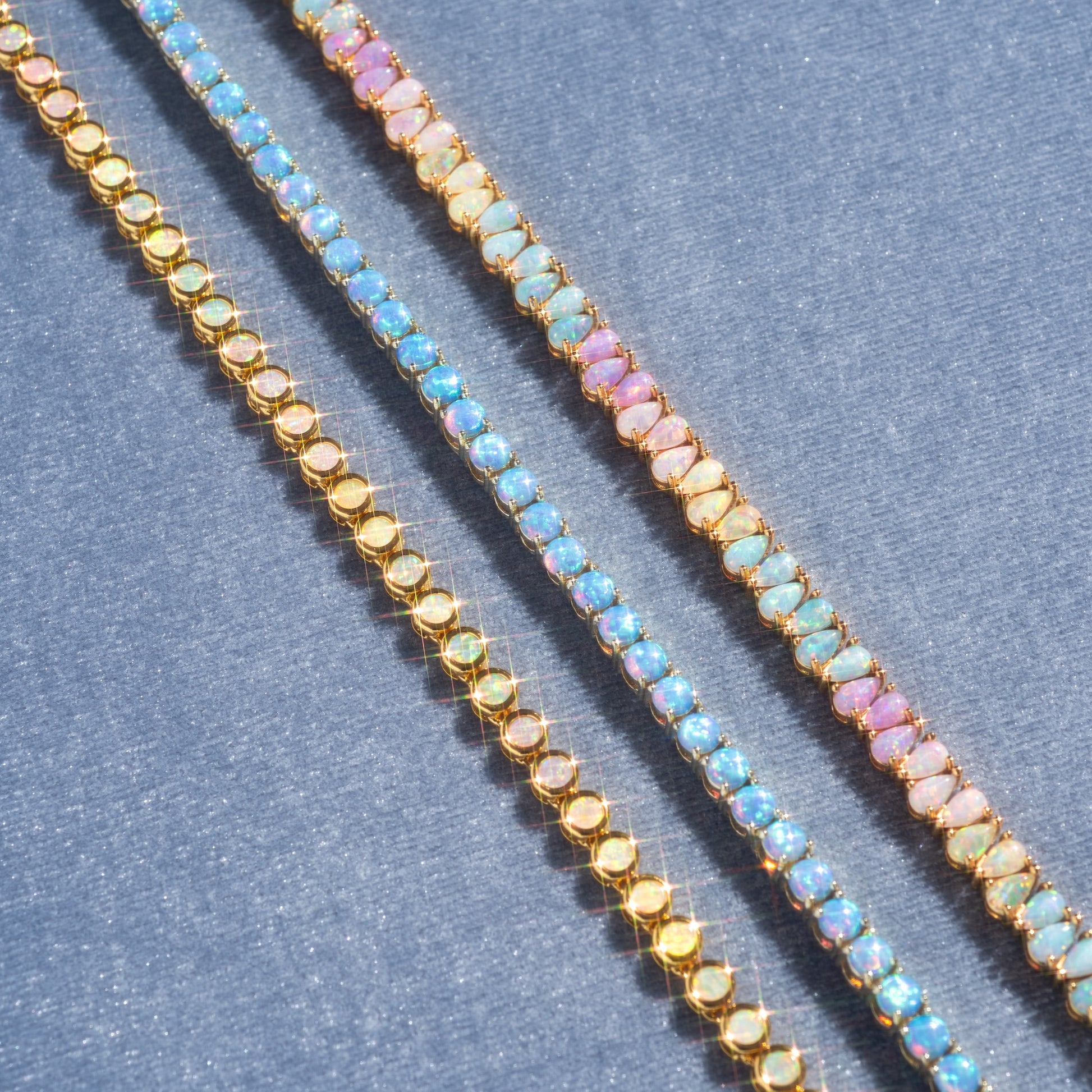 The Grand Heiress Tennis Necklace, featuring a repeating pattern of pastel multicolored gemstones in 18k gold-plated settings, is displayed diagonally on a textured blue surface.