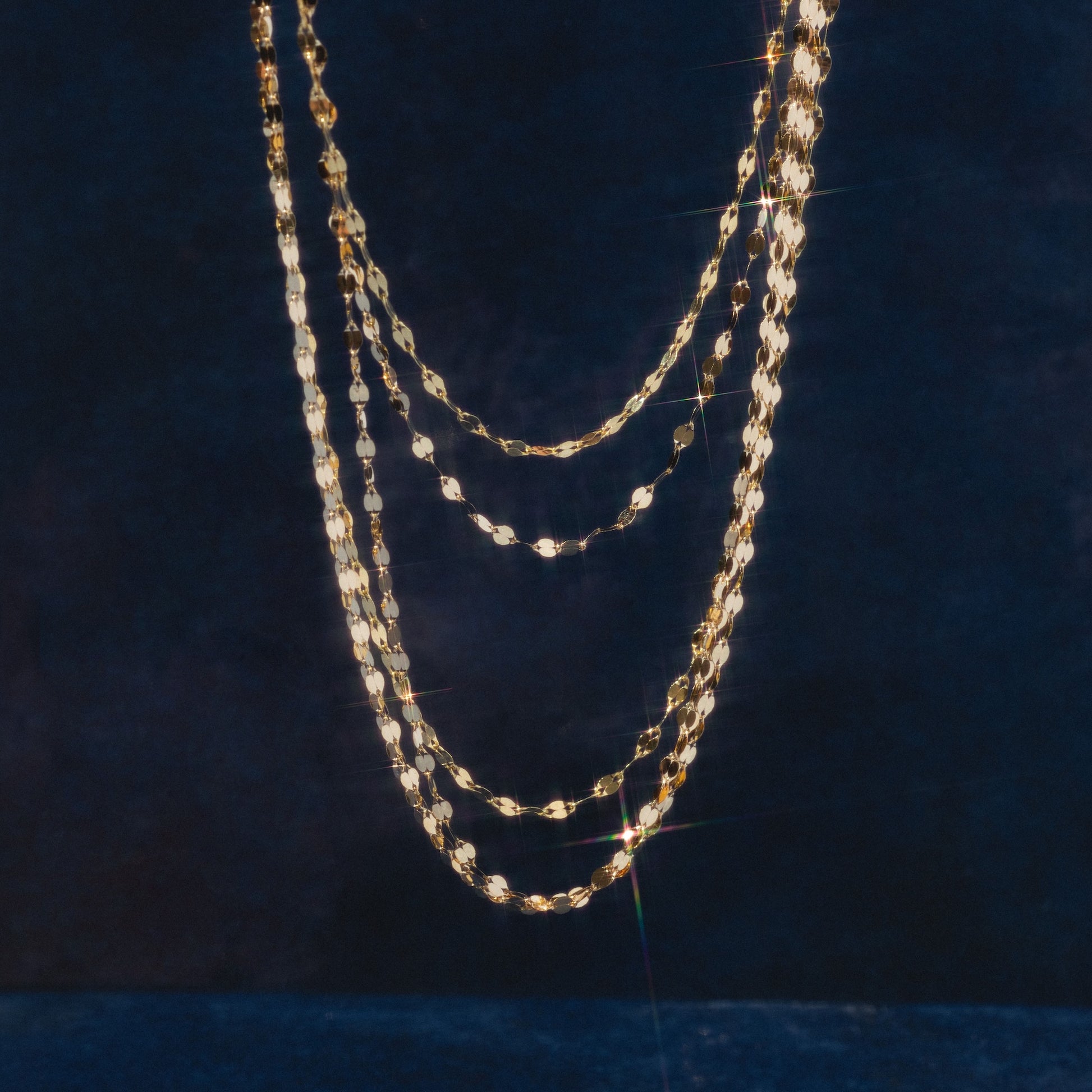 Four Solid Gold Italian Sparkle Link Chains in different lengths hang against a dark blue textured background, their surfaces glimmering as they catch the light and create radiant reflections.