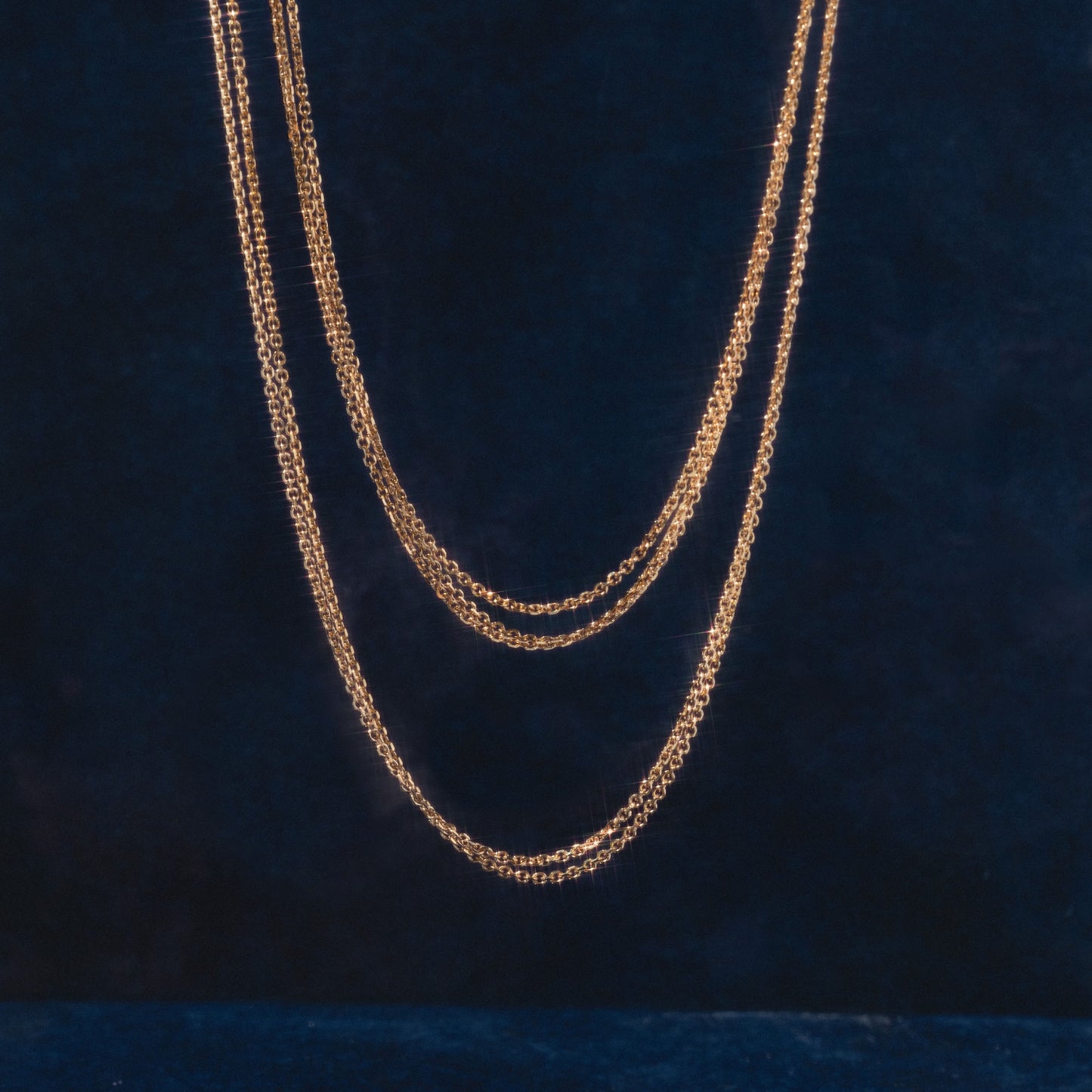 Three Solid Gold Simple Glimmer Gold Chains are elegantly layered in loops against a dark blue velvet background.