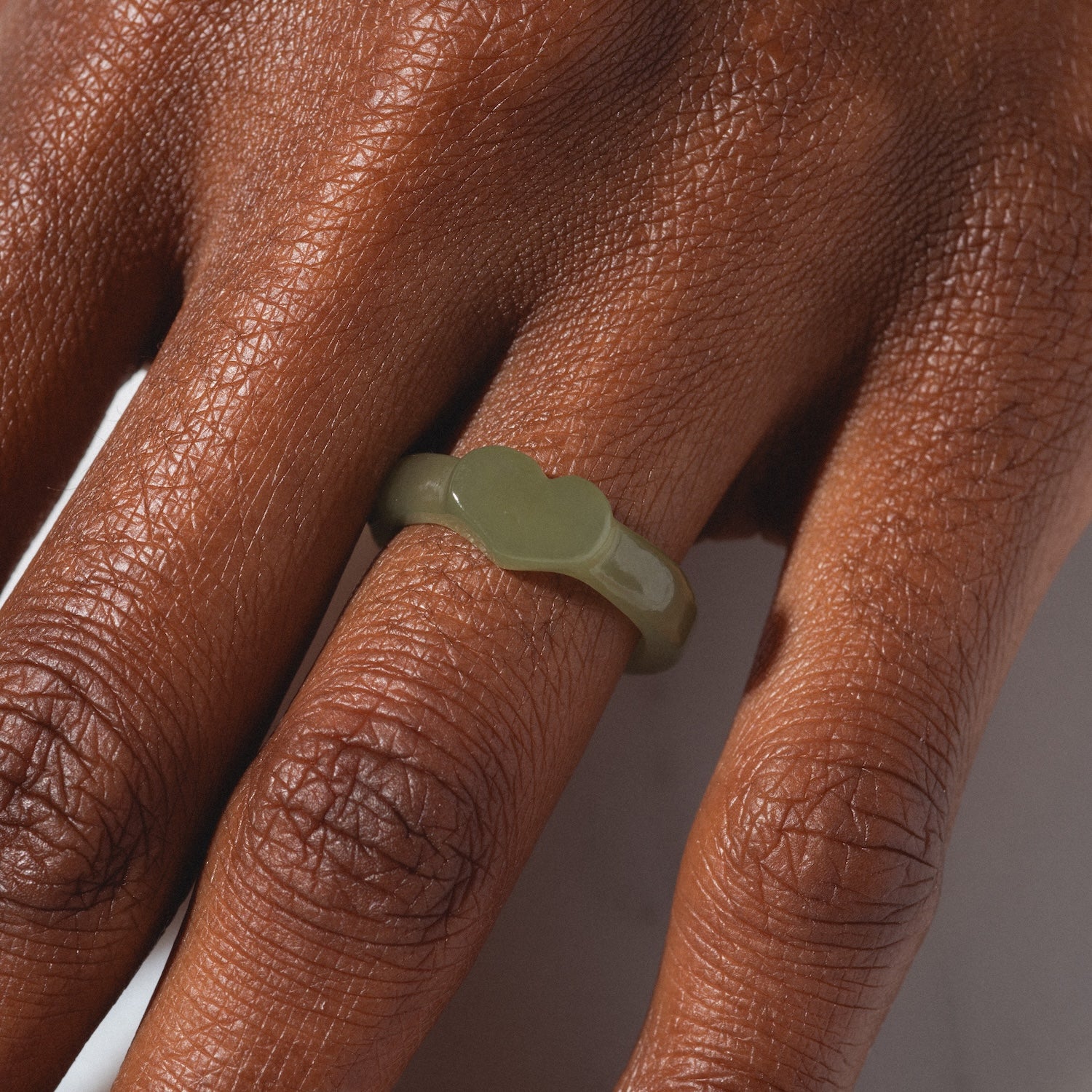 A close-up of a hand with dark skin wearing the Heart Jade Ring, a shiny, olive-green ring featuring a chunky, wavy design and a 3.5 mm band on the middle finger. The background is light and blurred.