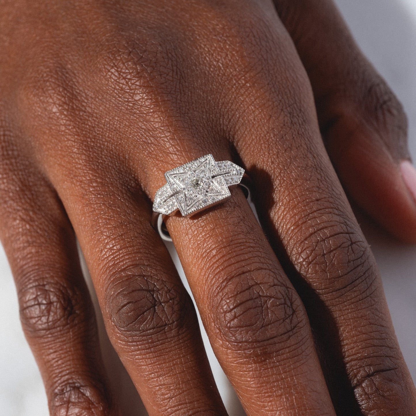 A close-up of a hand wearing the Art Deco Inspired Statement Ring, featuring a silver band with a central square-cut diamond and smaller diamonds in a geometric design.