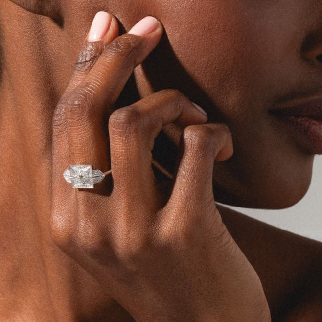 A woman with braided hair, wearing a gray tank top, looks to the side and showcases the Art Deco Inspired Statement Ring on her finger against a neutral background.