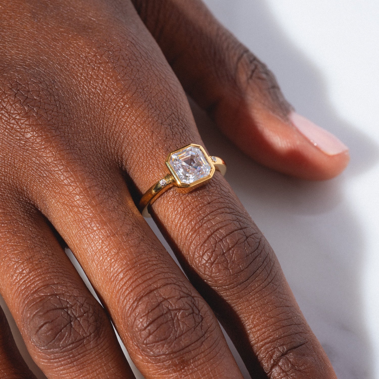 A close-up of a dark-skinned hand wearing the White Topaz Statement Star Set Band with a large, square-cut clear gemstone on the ring finger, light pink manicure, and a white surface in the background.