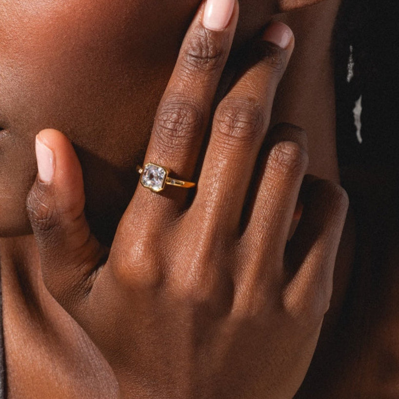 A woman with braided hair looks to the side, touching her face. She wears the White Topaz Statement Star Set Band on her ring finger and a gray tank top, against a softly lit background.