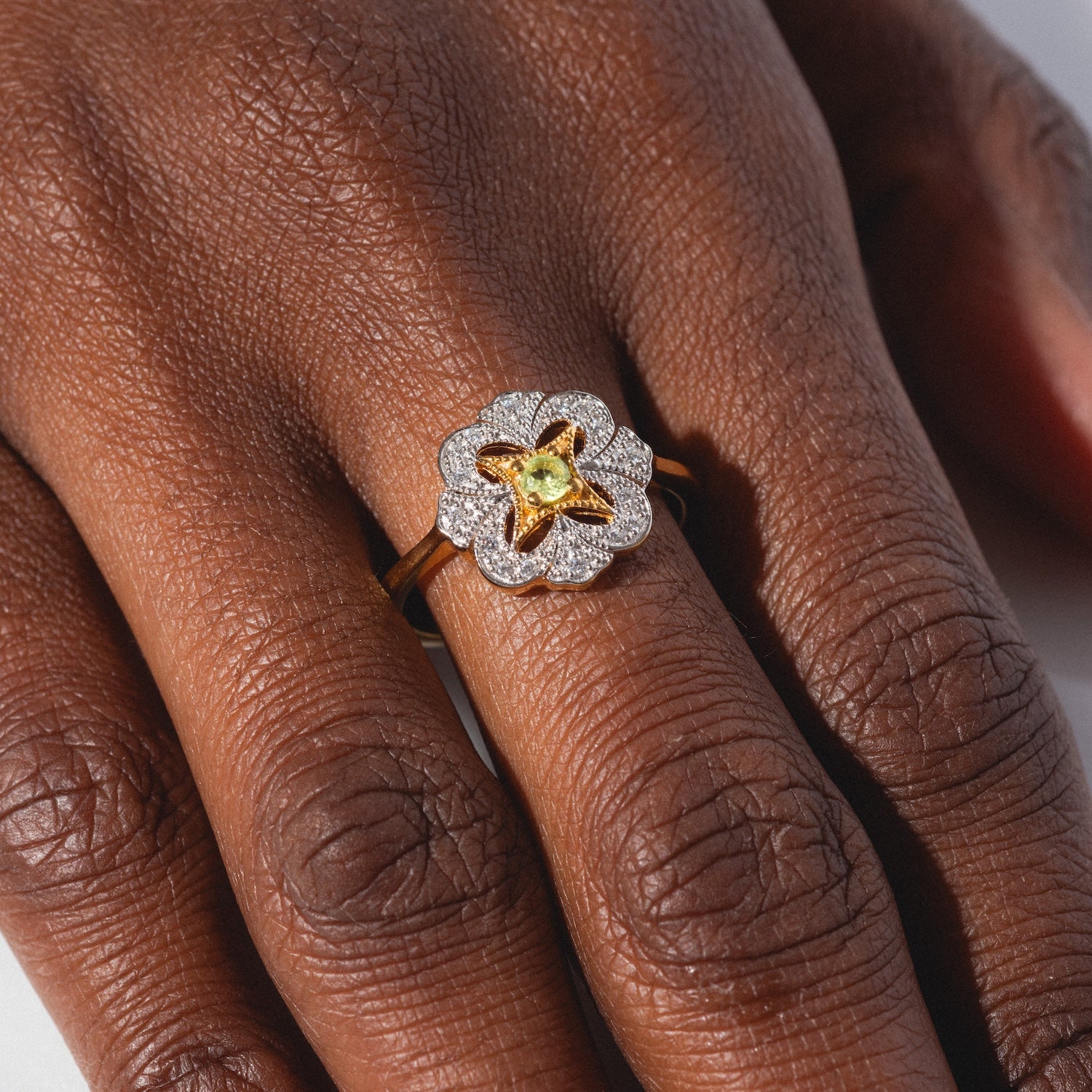 A close-up of a hand wearing the Floral Art Deco Inspired Peridot Ring, a gold ring with a silver flower-shaped design, showcasing a yellow peridot gemstone in the center and small diamonds outlining the petals.