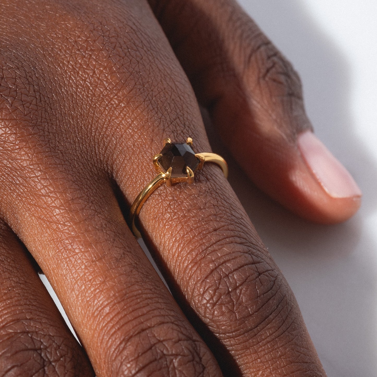 A close-up of a hand wearing the Hexagon Smokey Quartz Ring, featuring a gold band and a dark smokey quartz gemstone in a prong setting, resting on a light surface with soft lighting that highlights the ring and skin texture.