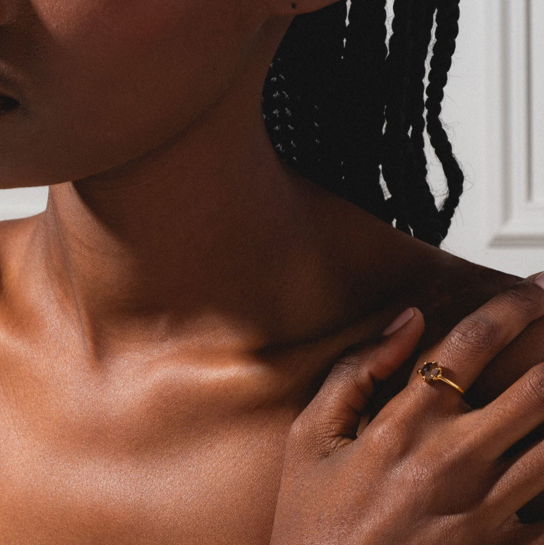 A close-up of a woman with braided hair touching her shoulder shows her wearing the Hexagon Smokey Quartz Ring. Sunlight casts shadows on her skin against a white wall background.