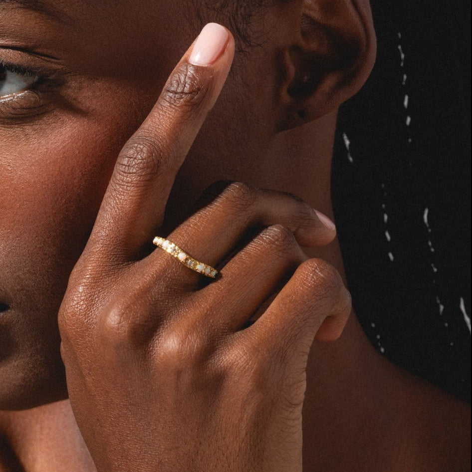 A woman with braided hair, dressed in a grey tank top and light pink nail polish, touches her face with her left hand. She appears thoughtful while wearing the Opal Stacking Wave Ring.