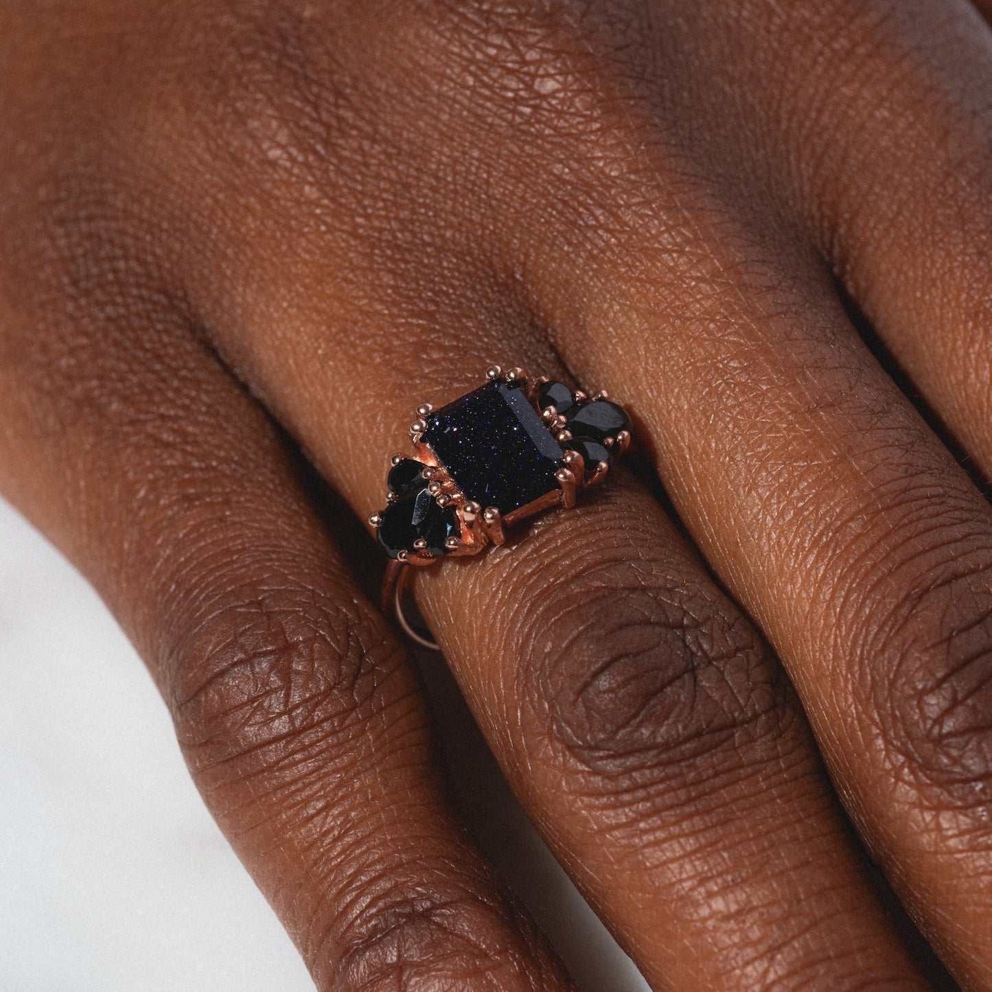 A close-up of a dark-skinned hand wearing the Gemstone Empress Ring, featuring a large rectangular dark blue stone and two smaller black stones, set in gold against a light background.