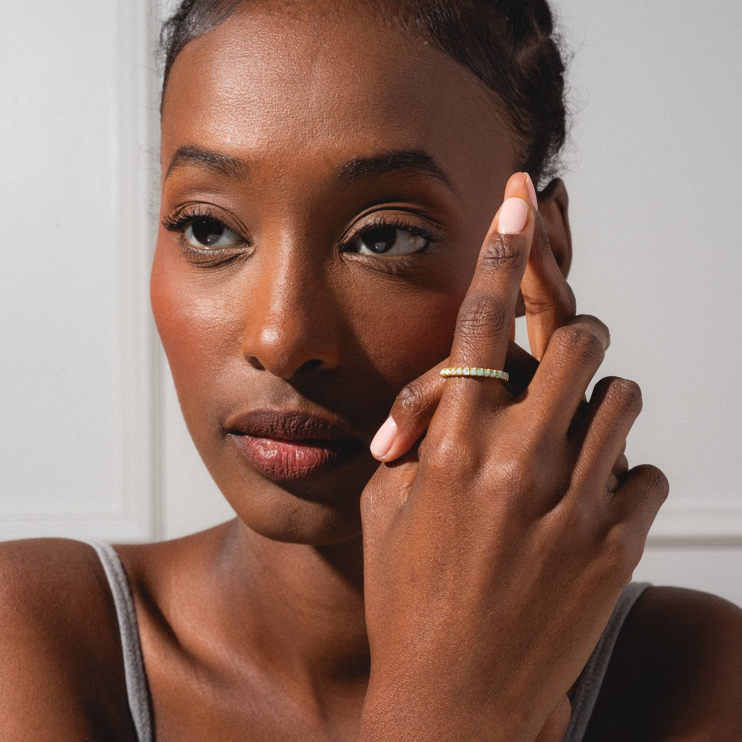 A woman with smooth brown skin and subtle makeup touches her face, elegantly showcasing the Indecisive Opal and Amazonite Eternity Band on her finger as she gazes thoughtfully into the distance in a gray tank top.