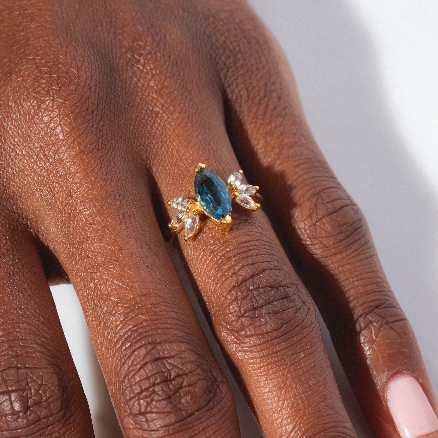 A close-up of a hand with neatly manicured, light pink nails wearing the Gemstone Firefly Ring—yellow gold vermeil with a large marquise-cut blue gemstone center and small clear side stones on the ring finger.