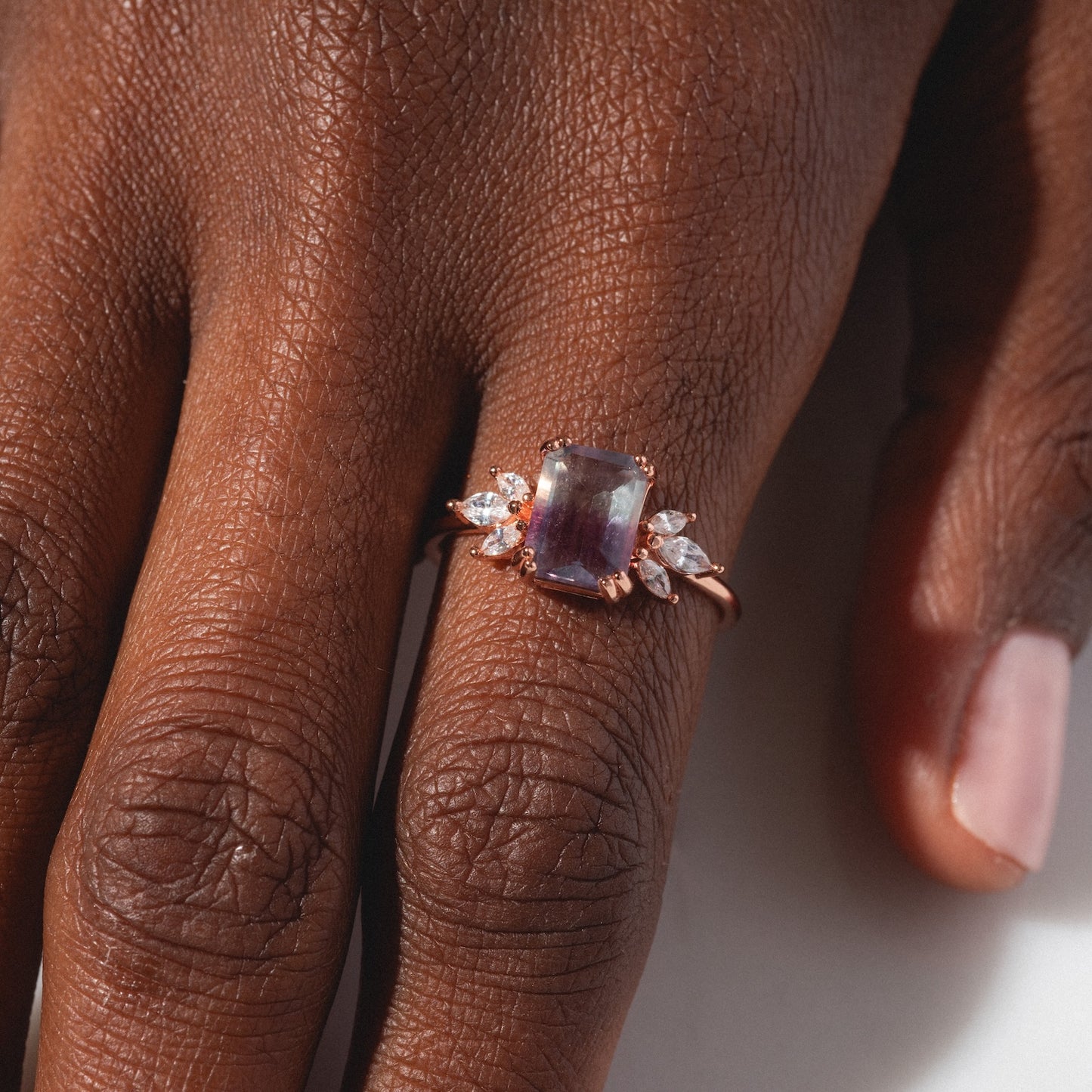 A close-up of a hand wearing the Rose Gold Fluorite Topaz Hazy Dream Ring, featuring a rectangular purple fluorite and small clear stones, set in rose gold vermeil, resting on a wooden surface.