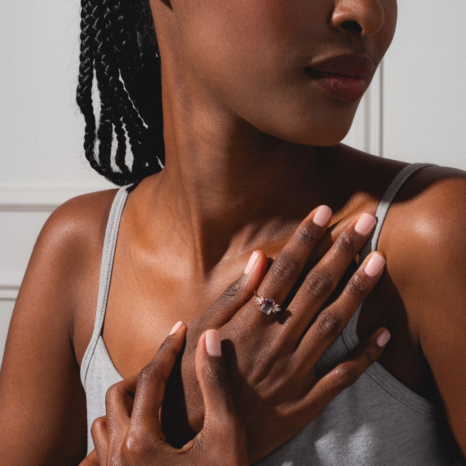 A woman in a gray camisole with braided hair touches her collarbone, showcasing the Rose Gold Fluorite Topaz Hazy Dream Ring. Manicured nails and soft lighting accentuate her glowing skin and the ring’s exclusive Local Eclectic design.
