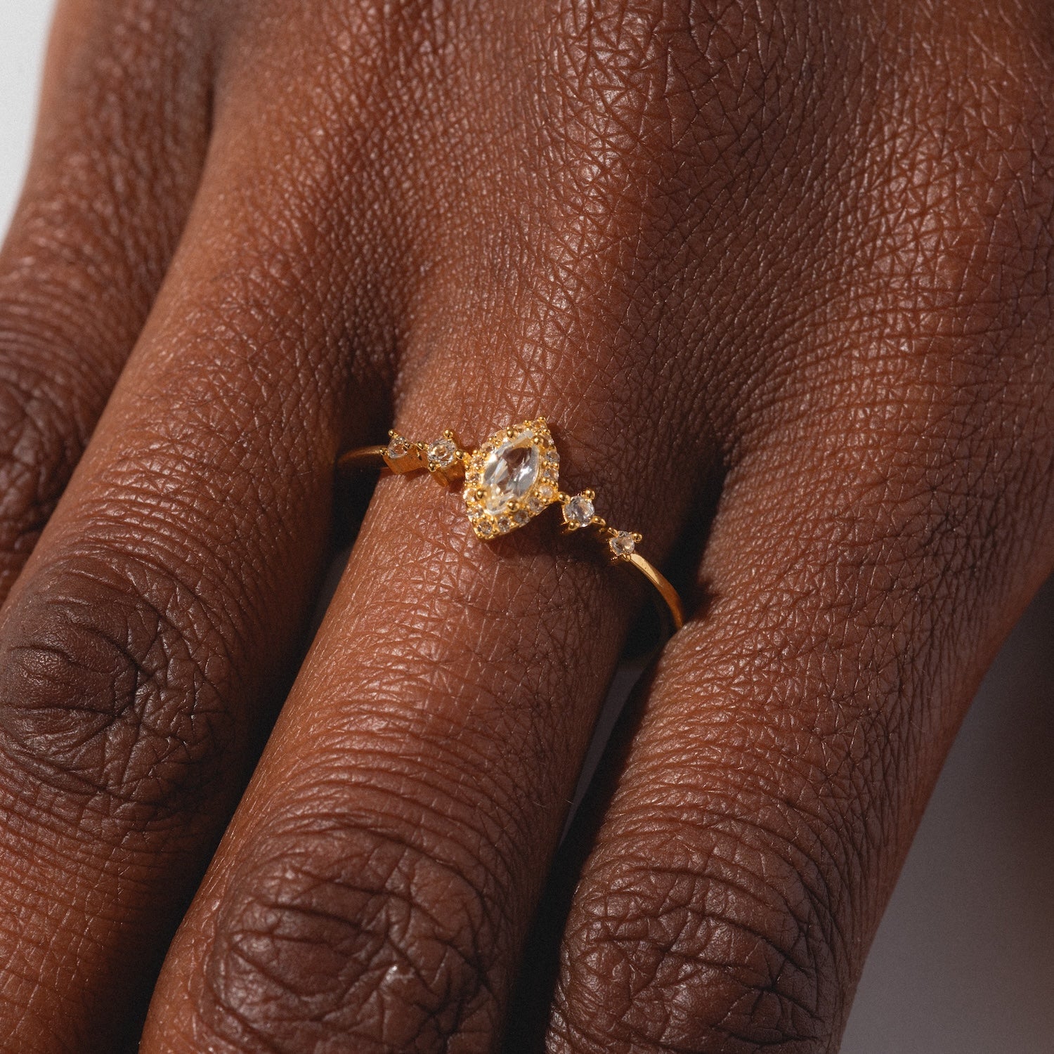 A close-up of a hand wearing the White Topaz Harlow Ring—an 18k gold vermeil marquise ring with a clear center gemstone and small round stones—set against a plain background.