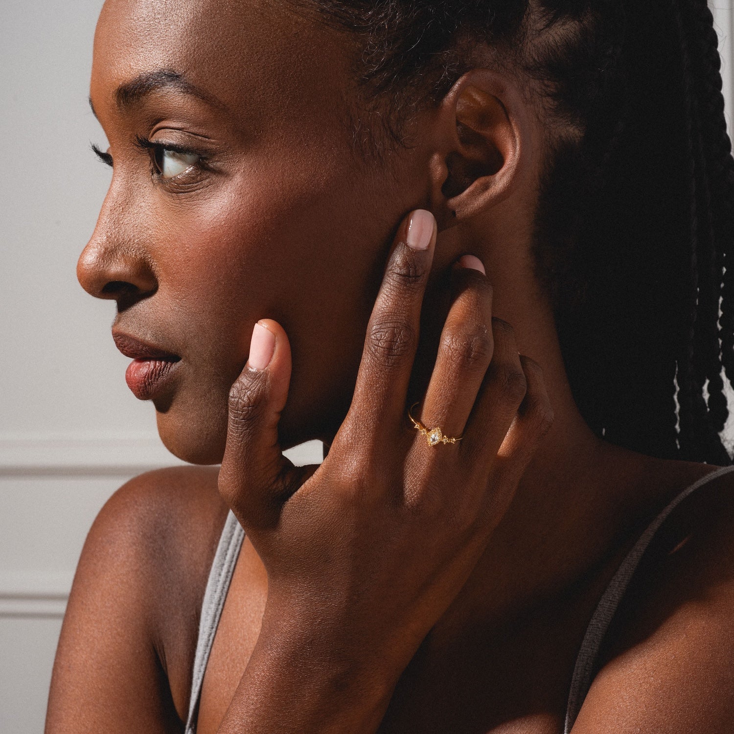 A woman with braided hair gazes left, touching her ear and face. She wears the White Topaz Harlow Ring in 18k gold vermeil, natural makeup, glowing skin, and a sleeveless top.