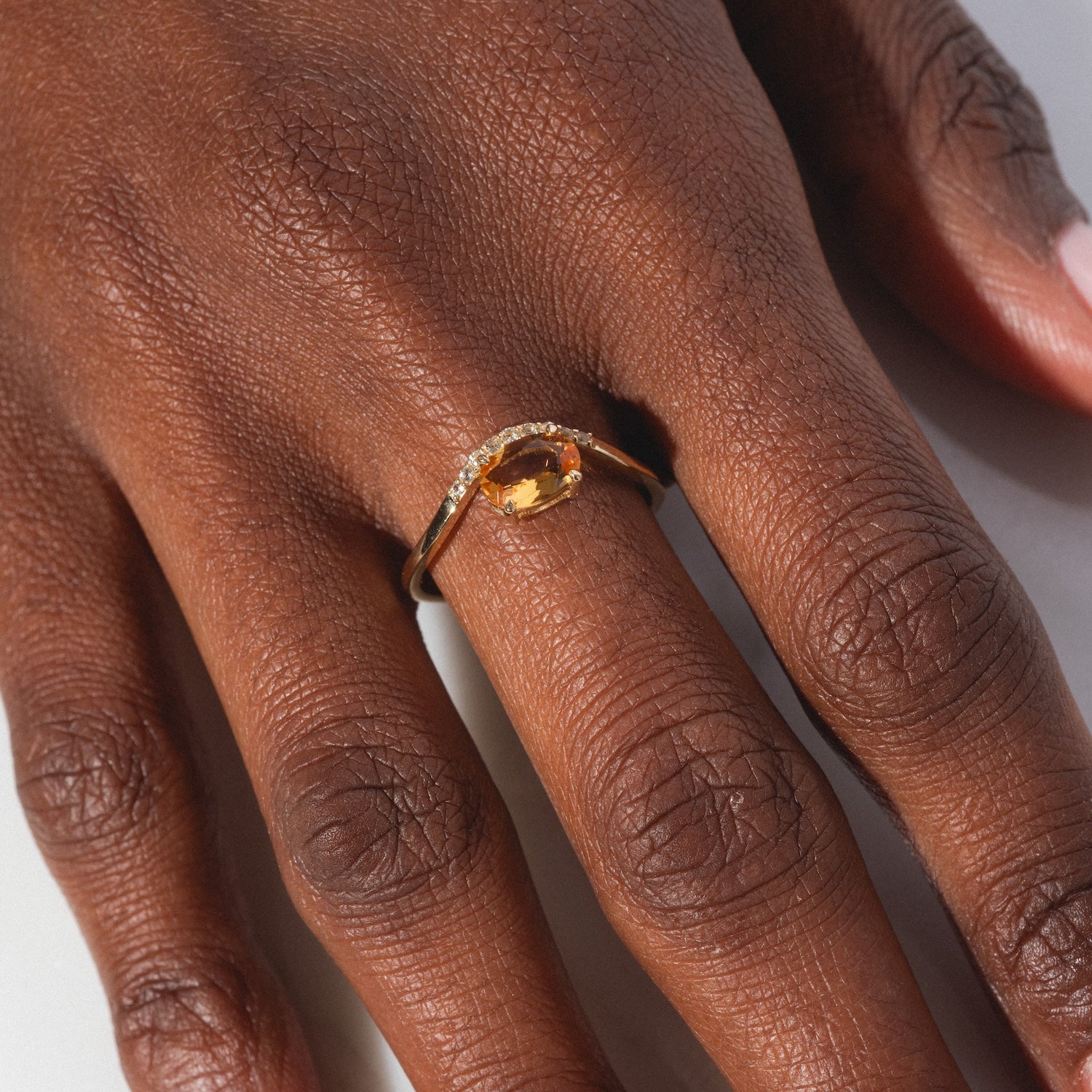 A close-up of a hand wearing the Solid Gold 2025 November Capsule Floating Citrine Statement Ring, featuring an oval citrine gemstone and small clear stones on the gold band, shown against a neutral background.