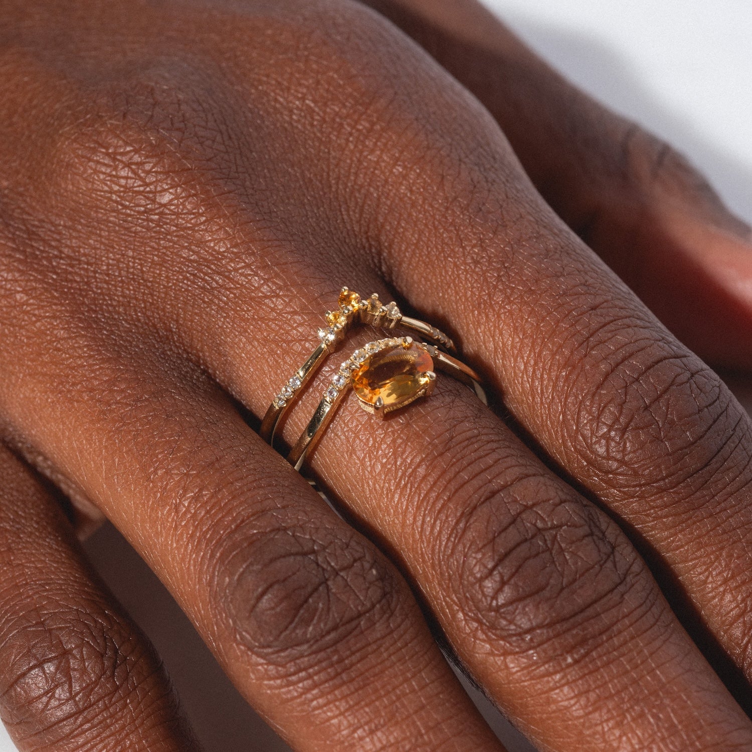A close-up of a dark-skinned hand wearing the Solid Gold 2025 November Capsule Citrine Ring Set, featuring a yellow citrine oval stone, sparkling white accents, and a delicate curved gold band. Free gift included.