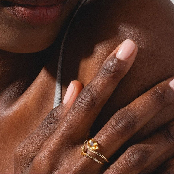 A woman with braided hair and natural makeup, wearing a gray tank top, rests her hand—adorned with the Solid Gold 2025 November Capsule Delicate Arch Citrine Stacking Ring—on her shoulder as she gazes thoughtfully to the side against a white paneled background.