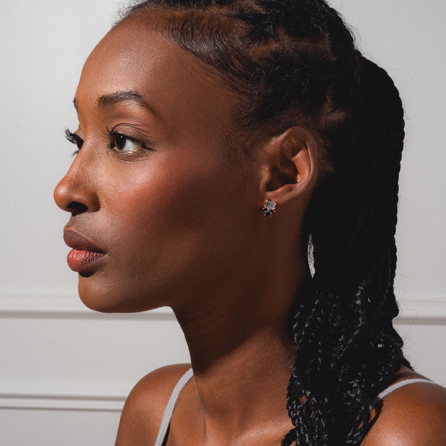 A woman with braided hair wears the Gemstone Empress Stud Earrings, delicate and flower-shaped, in profile against a plain background. Soft lighting highlights her smooth skin and light tank top.