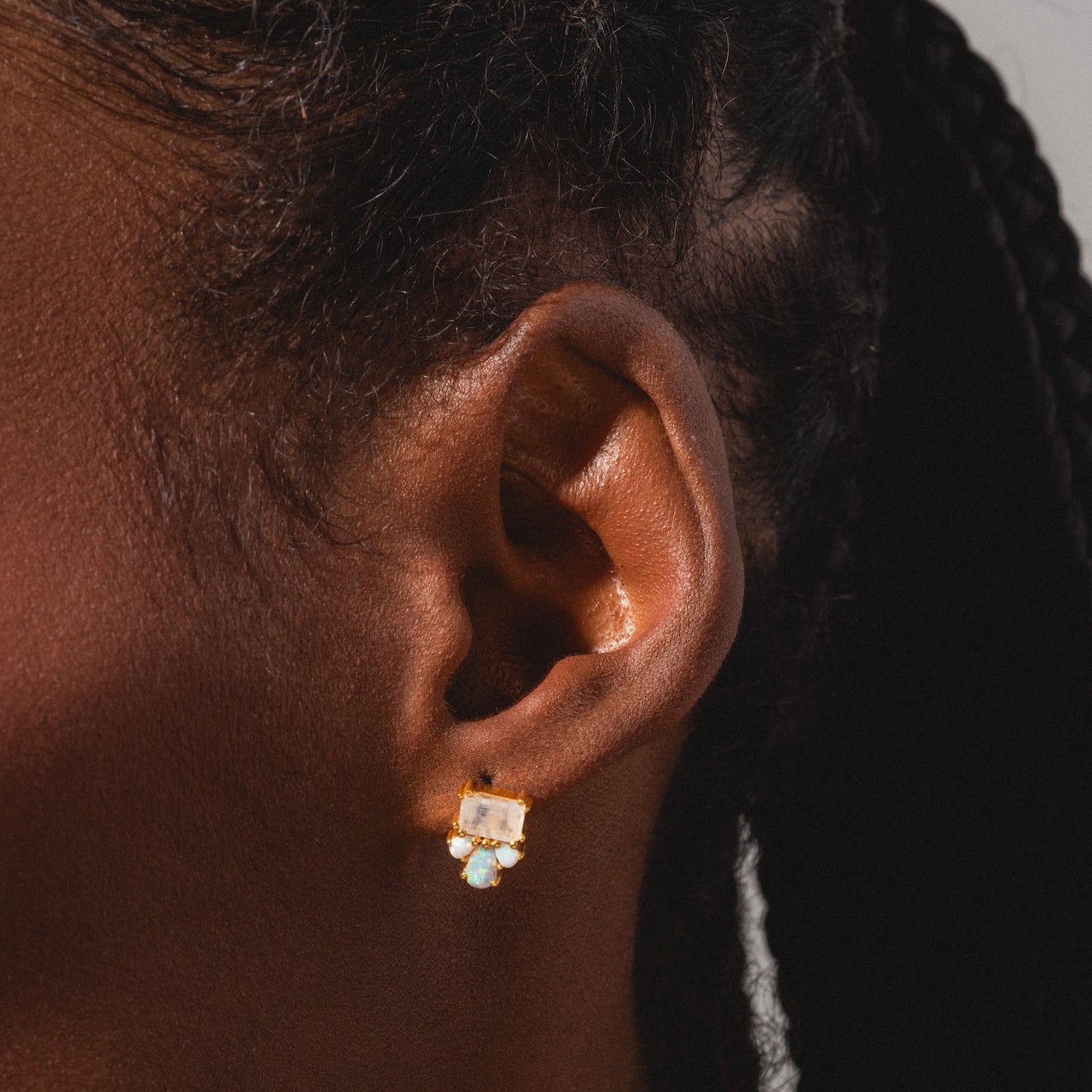 Close-up of a person with dark skin and braided hair pulled back, wearing Gemstone Empress Stud Earrings with a rectangular labradorite gemstone and a small round gem below.