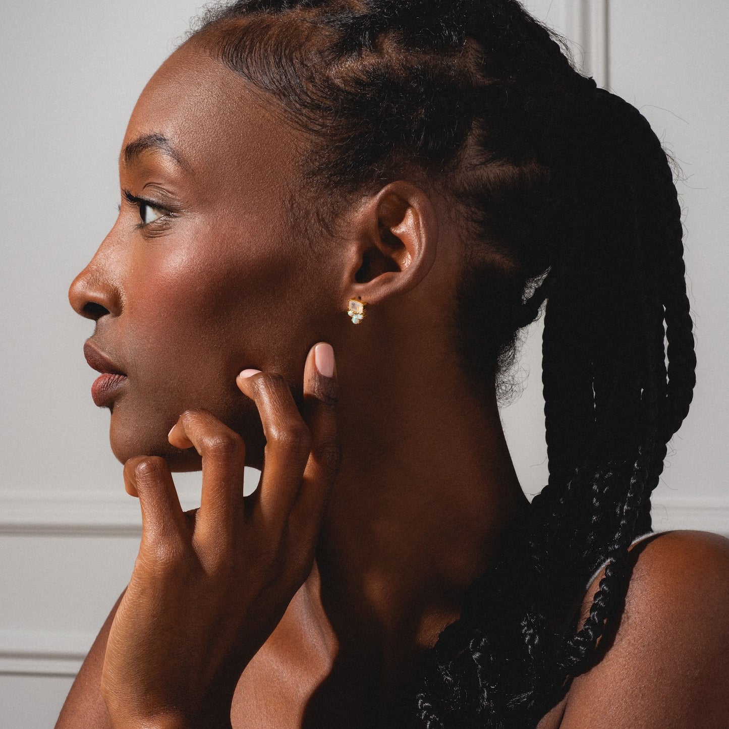 A woman with braided hair is shown in profile against a light background, resting her chin on her hand and wearing Gemstone Empress Stud Earrings shaped like flowers.