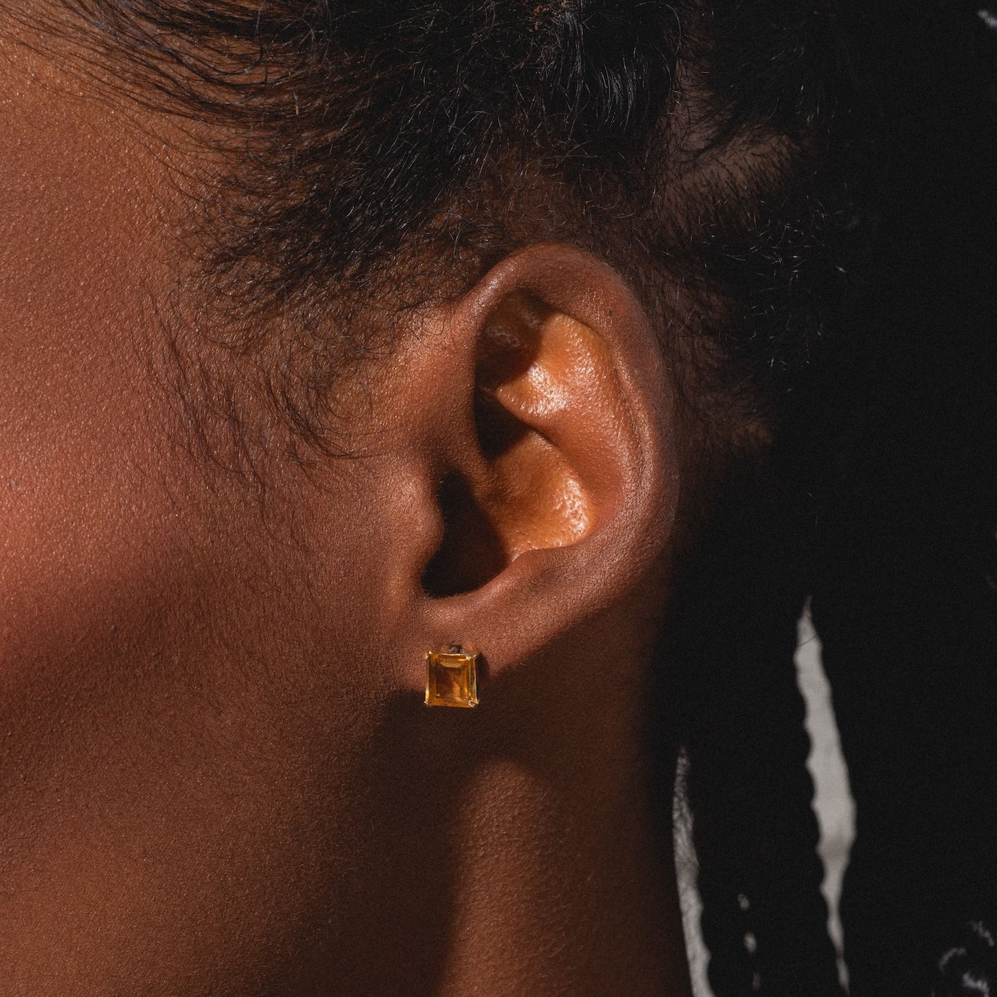 Close-up of a person's ear with dark skin and braided hair, wearing Solid Gold 2025 November Capsule Classic Citrine Stud Earrings—small, rectangular yellow gemstone studs.