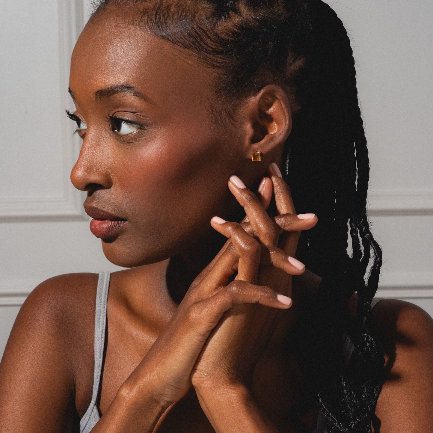 A woman with braided hair gazes left, hands near her face. She wears a gray tank top and Solid Gold 2025 November Capsule Classic Citrine Stud Earrings, their gleam subtly highlighted by soft natural light on her features.