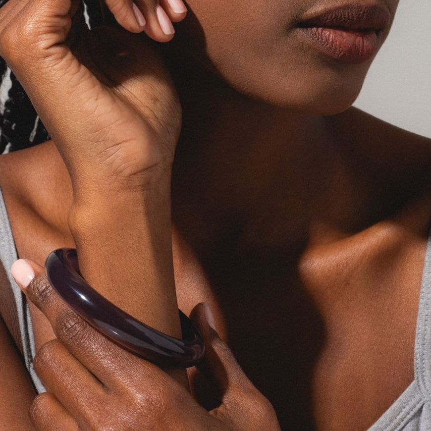 A woman with braided hair wearing a grey tank top rests her hand on her face, gazing thoughtfully to the side and showcasing the Moon Purple Jade Stone Bangle against a softly lit, neutral background.