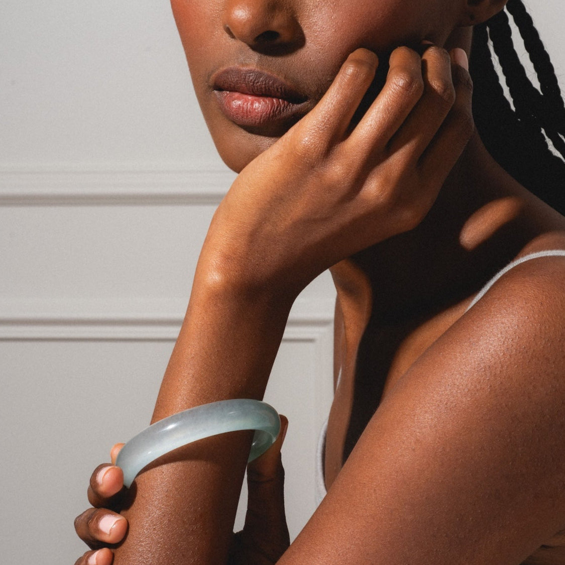 A woman with braided hair rests her chin on her hand, gazing to the side. She wears a grey tank top and a Pool Blue Jade Stone Bangle with a 2.4 inch opening and 13.5 mm width, softly lit across her face and arm.