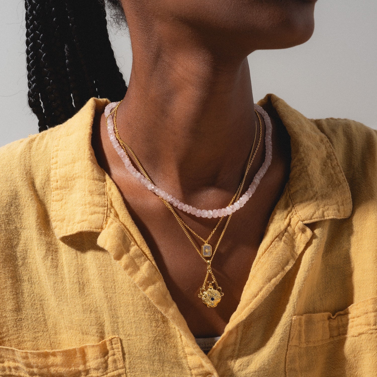 A person wearing a yellow textured shirt and three layered necklaces: a pink beaded necklace, a gold wishing star necklace with rectangular pendant, and the Rainbow Moonstone & Diamond Manifest Your Dreams Pendant Necklace.