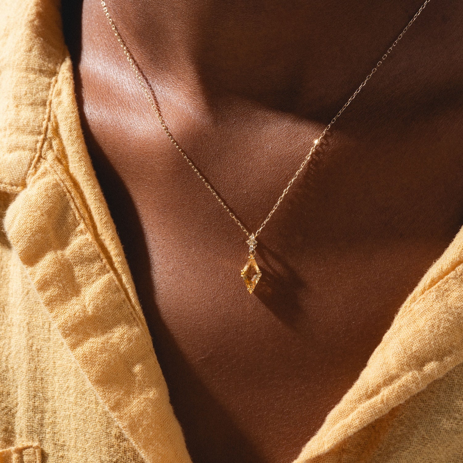A close-up shows a person wearing the Solid Gold 2025 November Capsule Art Deco Citrine Pendant Necklace over bare skin, paired with a yellow shirt. The geometric pendant hangs from a delicate chain.