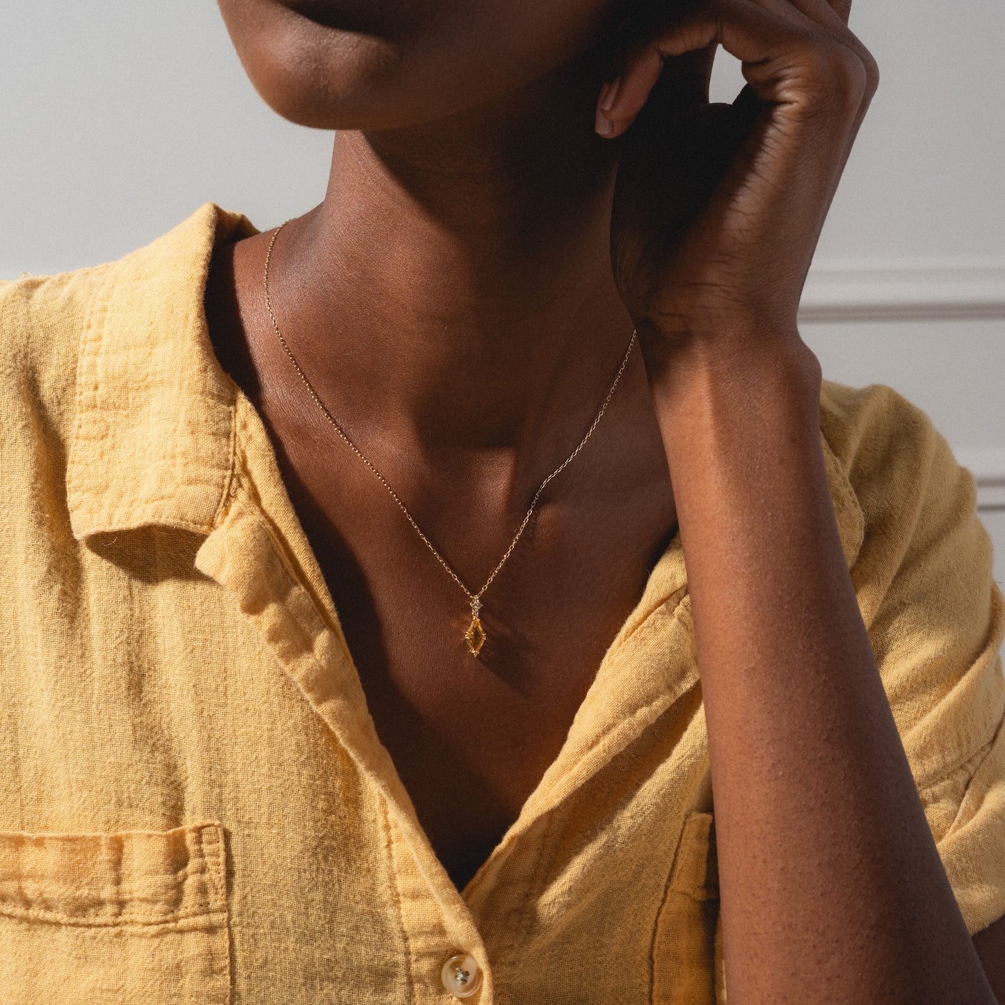 A person wears a textured yellow button-up and the Solid Gold 2025 November Capsule Art Deco Citrine Pendant Necklace, featuring a delicate geometric pendant, shown from shoulders to chin in soft natural light.
