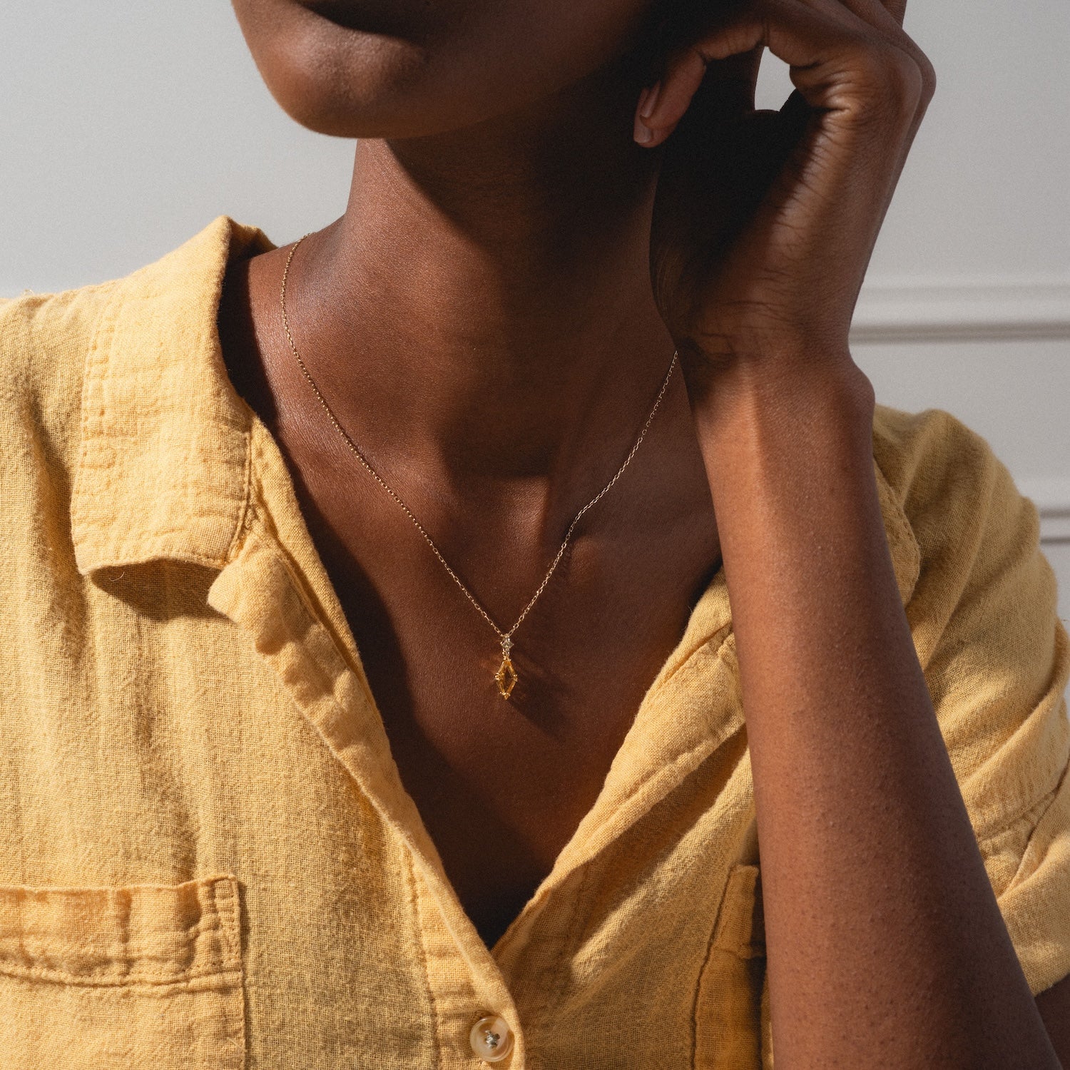 A person wears a textured yellow button-up and the Solid Gold 2025 November Capsule Art Deco Citrine Pendant Necklace, featuring a delicate geometric pendant, shown from shoulders to chin in soft natural light.