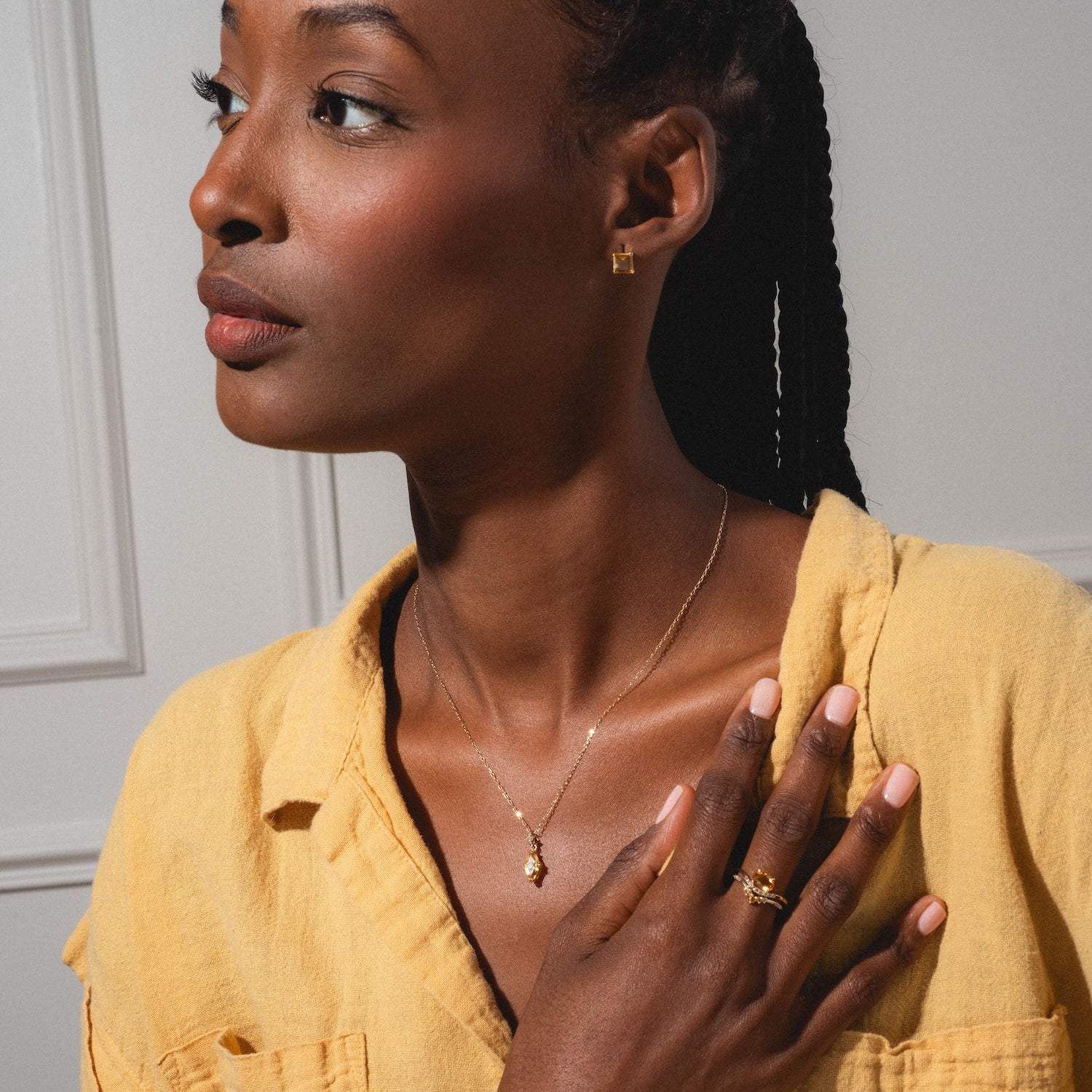 A woman with braided hair wears the Solid Gold 2025 November Capsule Citrine Full Set, featuring a gold necklace, earrings, and rings, as she poses against a light background with one hand resting on her chest. Free gift included.