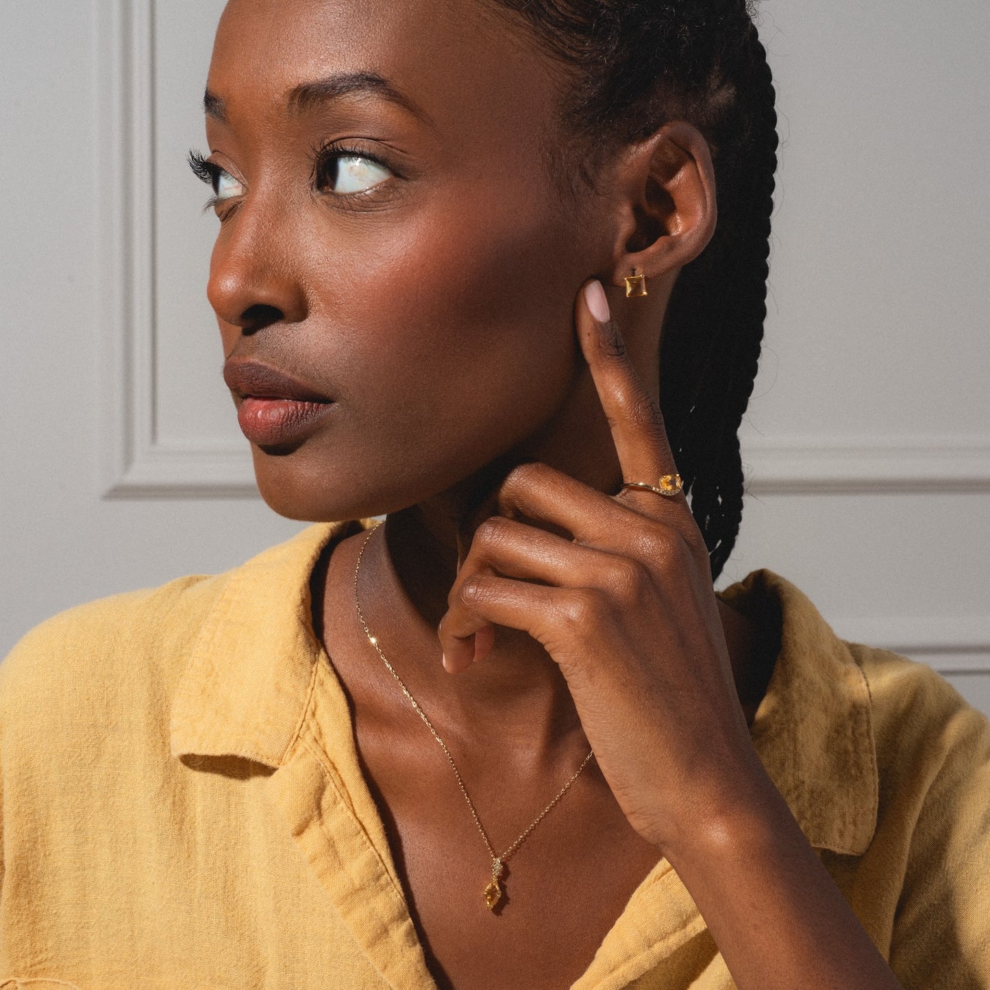 A woman in a yellow shirt poses against a light-paneled wall, touching her ear as she models the Solid Gold 2025 November Capsule Citrine Statement Set, including necklace, ring, and earrings—plus a free gift.