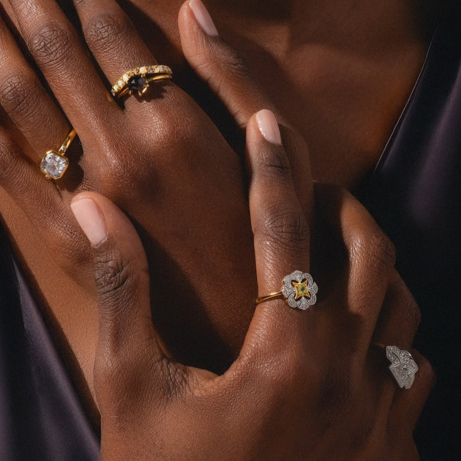 A close-up of hands with manicured nails wearing vintage-inspired jewelry, featuring the Hexagon Smokey Quartz Ring alongside a solitaire and textured band, set against a dark fabric background.