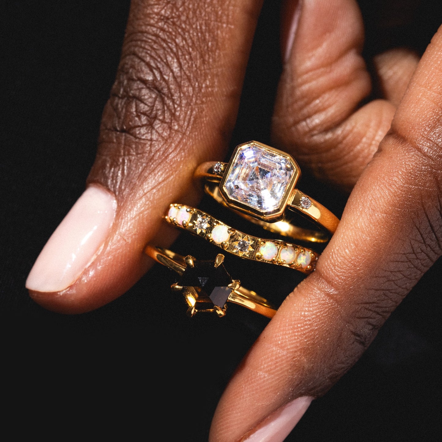 A hand with light pink nail polish wears three gold rings, including a 14k gold vermeil vintage-inspired band, a bold black star-shaped gemstone ring, and the shimmering Hexagon Smokey Quartz Ring.