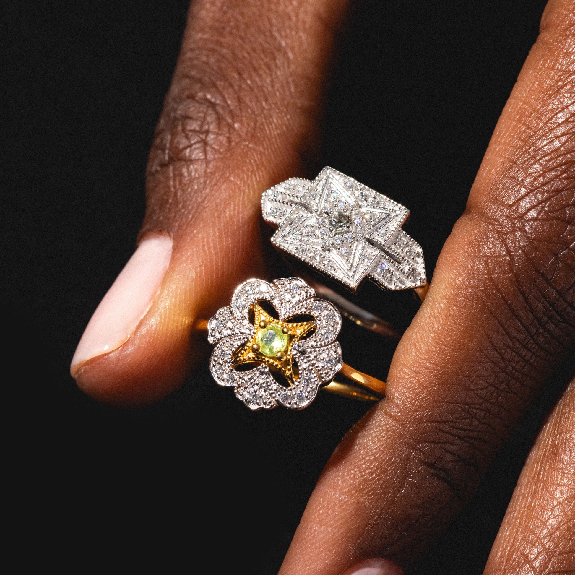 A hand with dark skin holds two ornate rings: a vintage-inspired silver band with a square diamond, and the Floral Art Deco Inspired Peridot Ring in 14k yellow gold vermeil with a flower-shaped design, set against a black background.