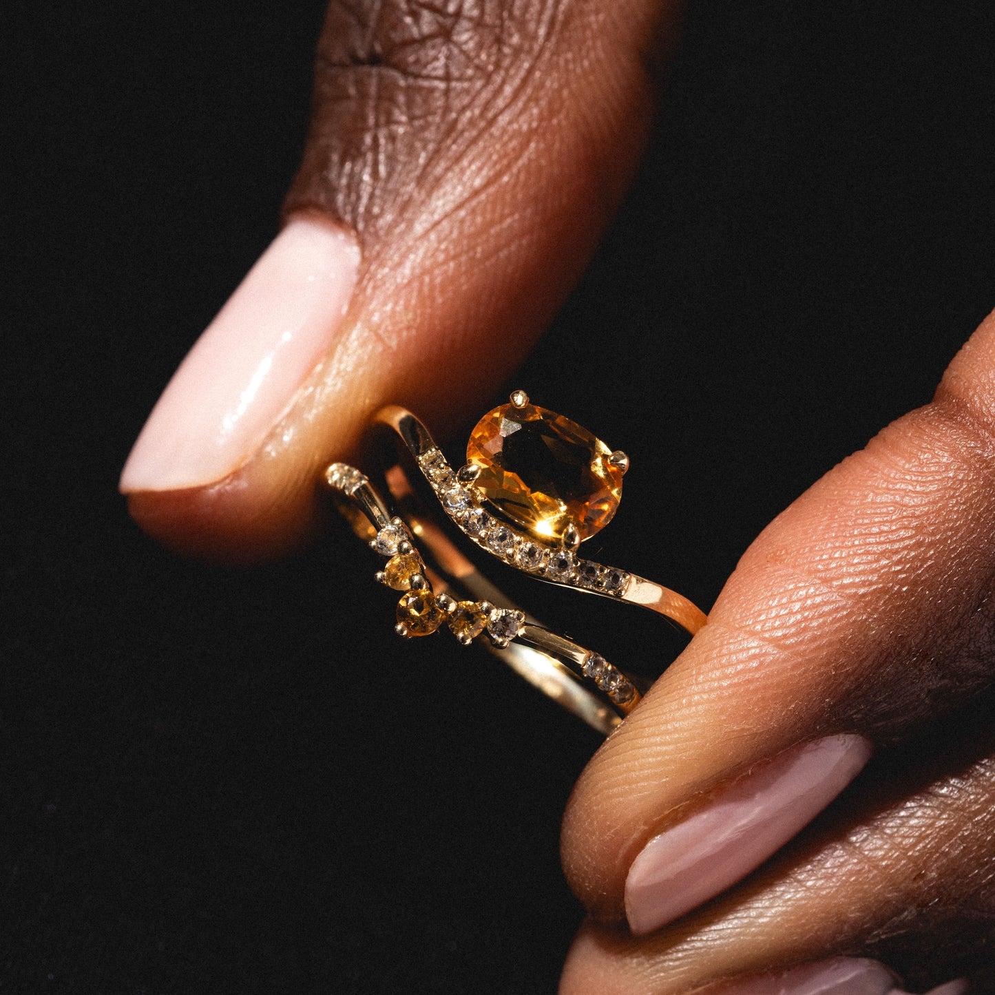 A hand with light pink nails holds the Solid Gold 2025 November Capsule Delicate Arch Citrine Stacking Ring, featuring a large oval yellow citrine and small diamonds, against a black background.