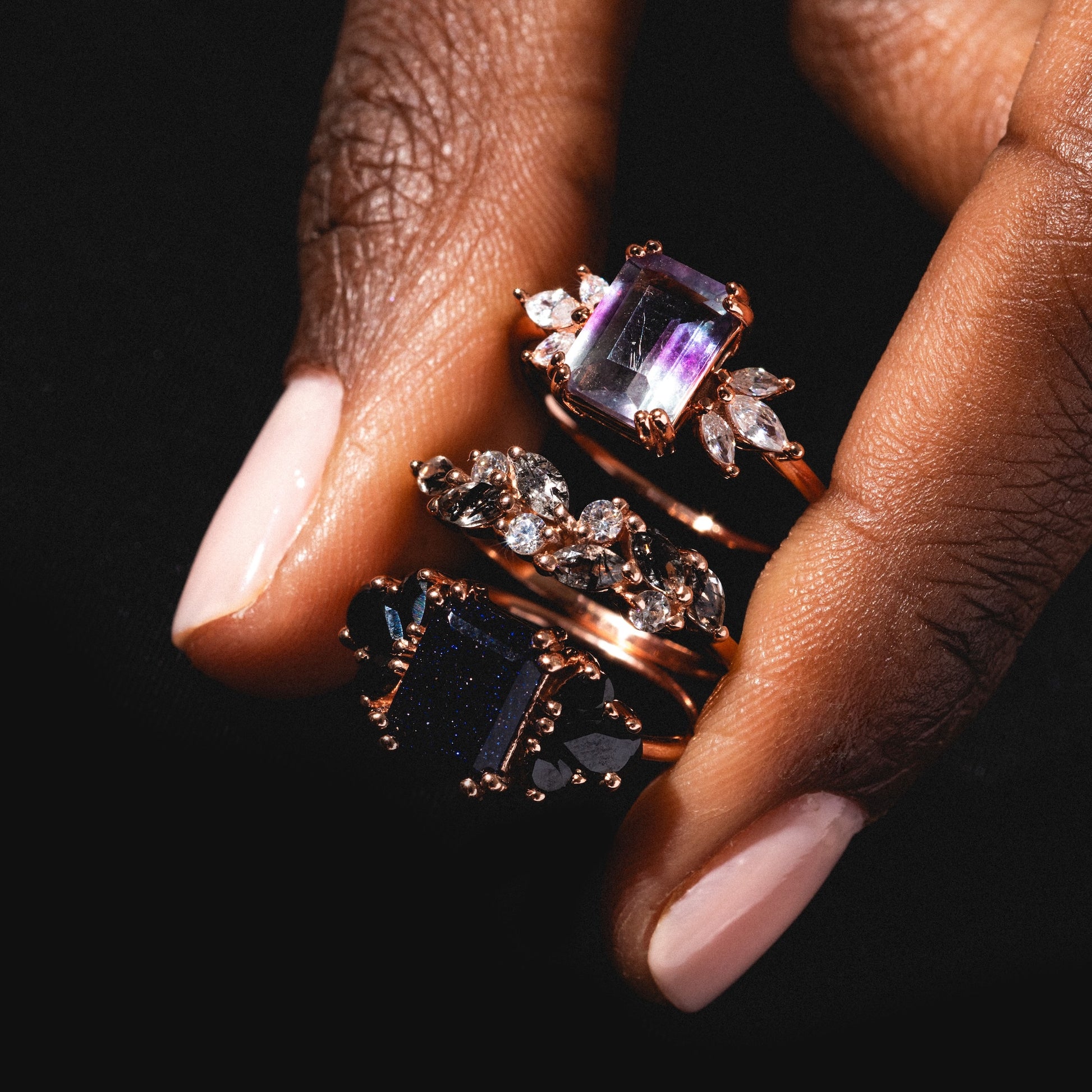 A hand with light pink nails displays three Gemstone Orchard Rings, each featuring a large gemstone—one with moss agate—surrounded by smaller sparkling stones, set against a dark background.