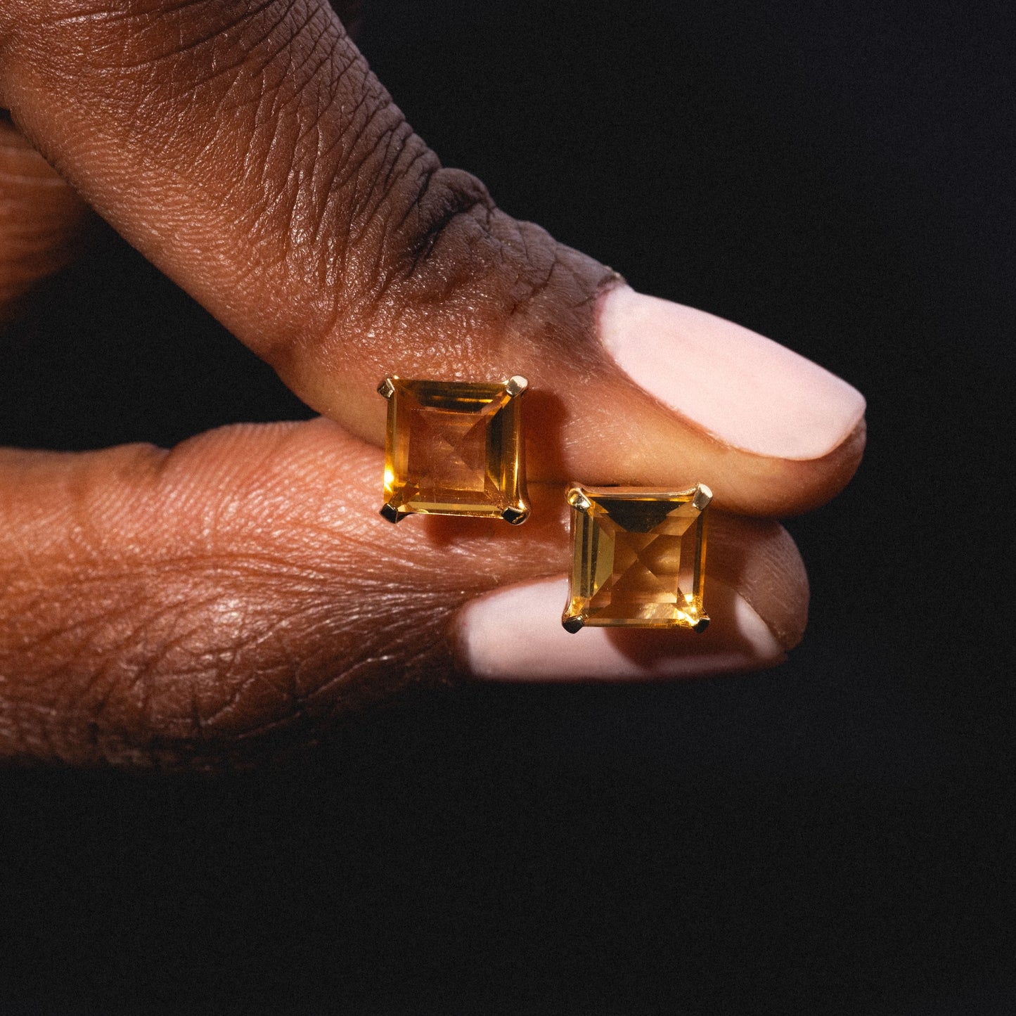 A hand with light pink nail polish holds two Solid Gold 2025 November Capsule Classic Citrine Stud Earrings against a dark background.