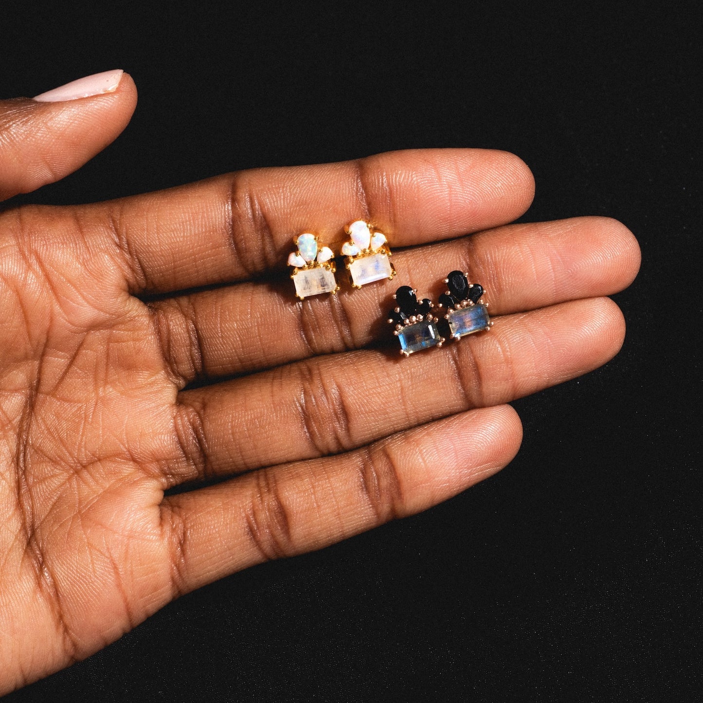 A hand holds three pairs of Gemstone Empress Stud Earrings, each with rectangular colored gemstones and small round gems above, set in metal, against a black background.
