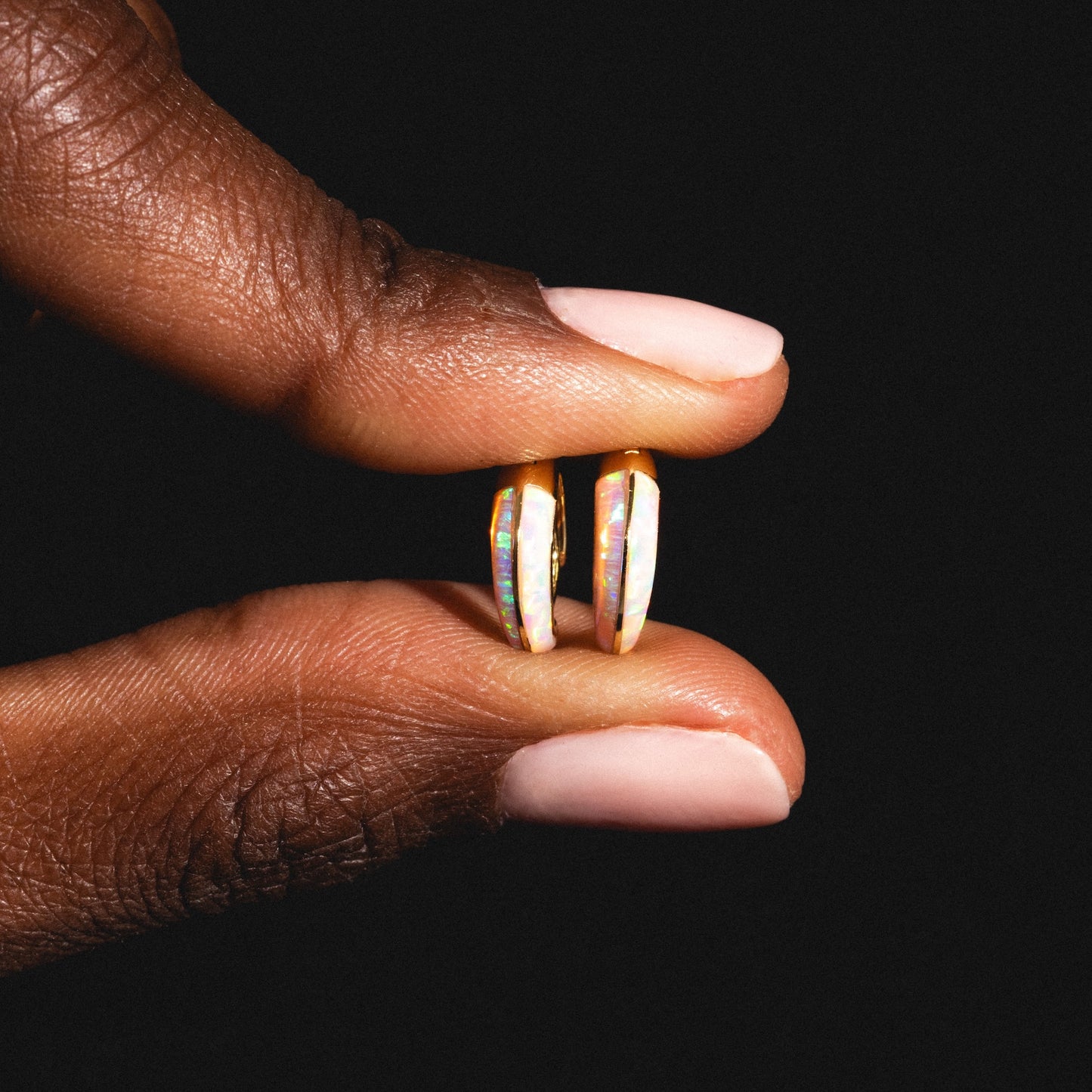 A hand with light pink nails holds the Opal Ombre Deux Huggies, crafted in gold vermeil, against a black background.