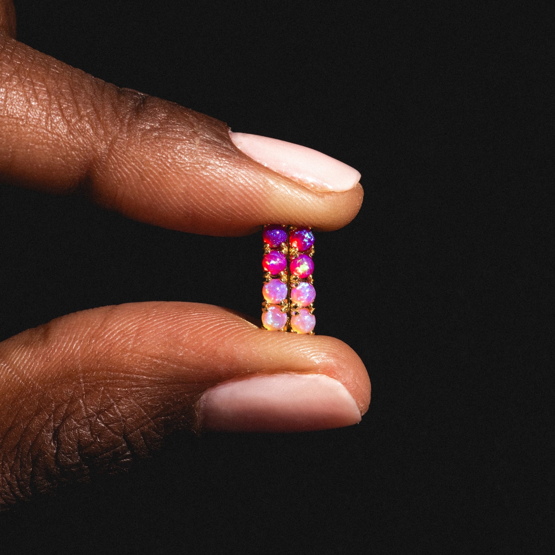 A hand with light pink polished nails holds the Pink Opal Ombre Oblong Ella Huggies—colorful earrings featuring red, pink, and purple lab-created opal beads—set against a striking black background.